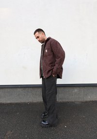 Veste marron texturée avec des éclats orange et bleu, associée à un pantalon foncé et des chaussures noires épaisses, debout contre un mur clair.