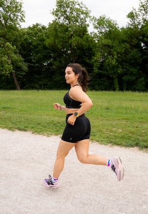 Top sportivo nero e pantaloncini, che corre su un sentiero di ghiaia. Scarpe da corsa viola e rosa. Erba verde e alberi sullo sfondo.