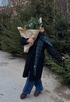 Abrigo negro, bufanda a cuadros verde y azul marino, sosteniendo un gran ramo de ramas perennes envueltas en papel kraft, con árboles y luces de fondo.
