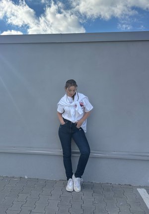 Camiseta blanca con diseño de nudo, jeans ajustados azul oscuro y zapatillas blancas. De pie contra una pared gris, con un cielo azul nublado arriba.