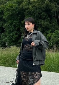 Veste en cuir noire portée sur une robe noire en dentelle transparente avec des motifs floraux, présentant un design superposé et des accents en chaîne argentée.
