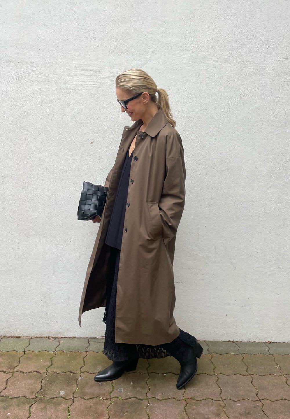 Brown trench coat featuring large buttons, side pockets, and a relaxed fit. It is worn over a black top and black skirt, complemented by black ankle boots.