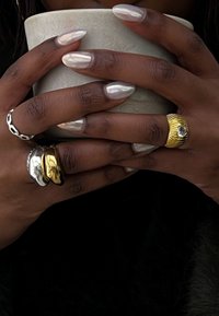 Keramiktasse, die von Händen mit langen, silberfarbenen Nagellack gehalten wird. Sie zeigt mehrere Ringe: einen silbernen Kettenring, einen goldenen und silbernen Ring sowie einen strukturierten goldenen Ring.