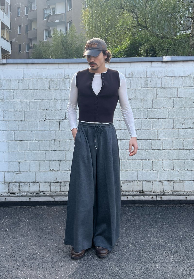 Homme portant une chemise blanche à manches longues sous une veste noire sans manches, un pantalon large gris, des chaussures marron et une casquette sombre, debout dehors près d’un mur en briques blanches.