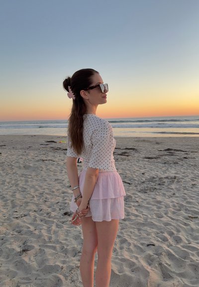 Mujer de cabello largo con una blusa blanca de lunares y falda rosa, de pie en una playa de arena al atardecer, mirando al océano con las manos entrelazadas detrás de la espalda.