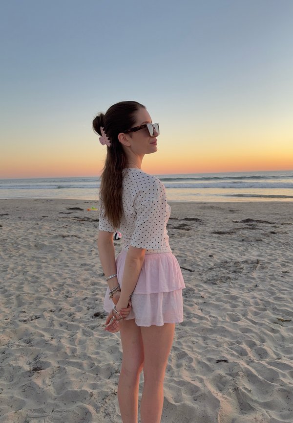 Mujer de cabello largo con una blusa blanca de lunares y falda rosa, de pie en una playa de arena al atardecer, mirando al océano con las manos entrelazadas detrás de la espalda.
