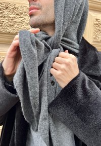 Écharpe en laine grise enroulée autour du cou, associée à un manteau sombre. Texture douce avec une finition lisse et des plis subtils visibles.