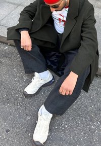 Άνδρας καθισμένος σε πεζοδρόμιο, φορώντας σκούρο πράσινο παλτό, κόκκινο σκουφάκι, γυαλιά ηλίου, παντελόνια με σχέδια, λευκά αθλητικά παπούτσια και ακουστικά.