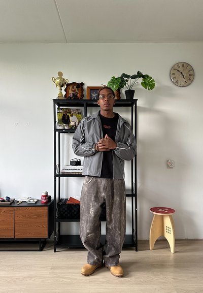 Chaqueta gris con cremallera y acentos blancos, camiseta negra, pantalones anchos de pata ancha con manchas marrones y zuecos de madera. Ambiente interior con estanterías y plantas.