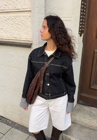 Črna jeans jakna z sivimi manšetami, bela majica in bele kratke hlače. Rjav usnjen torbica preko telesa. Dolgi, kodrasti lasje. Stoji ob teksturirani steni.