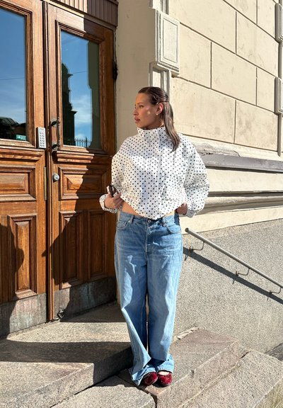 Mujer con chaqueta blanca de lunares y vaqueros azules está al aire libre junto a una puerta de madera y escalones de piedra, sosteniendo un teléfono y mirando hacia la derecha.