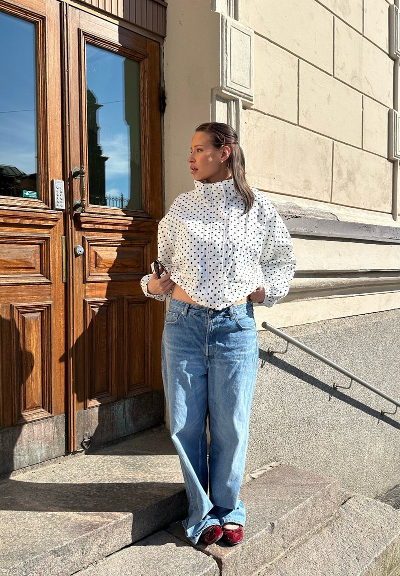 Woman in white polka dot jacket and blue jeans stands outdoors by wooden door and stone steps, holding phone and looking right.
