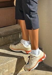 Ténis de corrida leves em bege com detalhes em laranja, parte superior texturizada, atacadores pretos e sola almofadada, combinados com meias brancas de tornozelo.