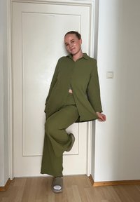 Conjunto de duas peças verde-oliva composto por uma camisa de botão de mangas longas e calças de perna larga. Design simples, textura suave, com calçado cinza.