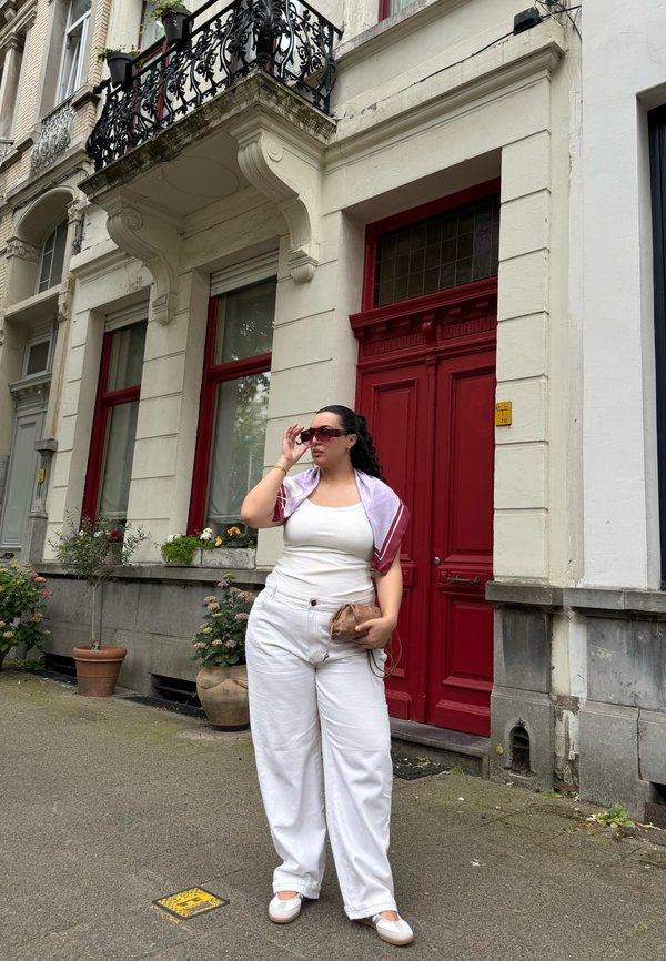 Femme en tenue blanche ajustant ses lunettes de soleil, debout sur le trottoir devant un bâtiment avec porte et encadrements de fenêtres rouges.