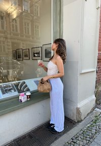 Γυναίκα με λευκή, ατμοσφαιρική τοπ χωρίς τιράντες και μπλε ρίγες παντελόνι, κρατώ�ντας μία καφέ τσάντα και μία κούπα καφέ, παρατηρεί μια βιτρίνα καταστήματος.