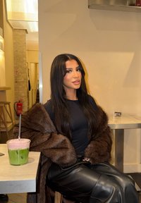 Un manteau en fausse fourrure marron est posé sur un haut noir ajusté, associé à un pantalon en cuir noir. Une boisson rose et verte est posée sur une table en bois clair.