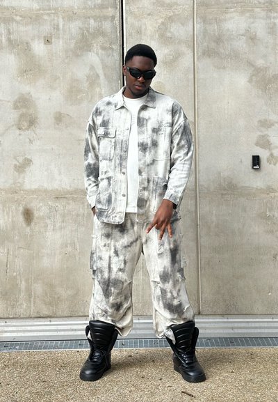 Chaqueta y pantalones sueltos de tie-dye en gris y blanco, combinados con botas gruesas negras. El look presenta múltiples bolsillos y un acabado texturizado.