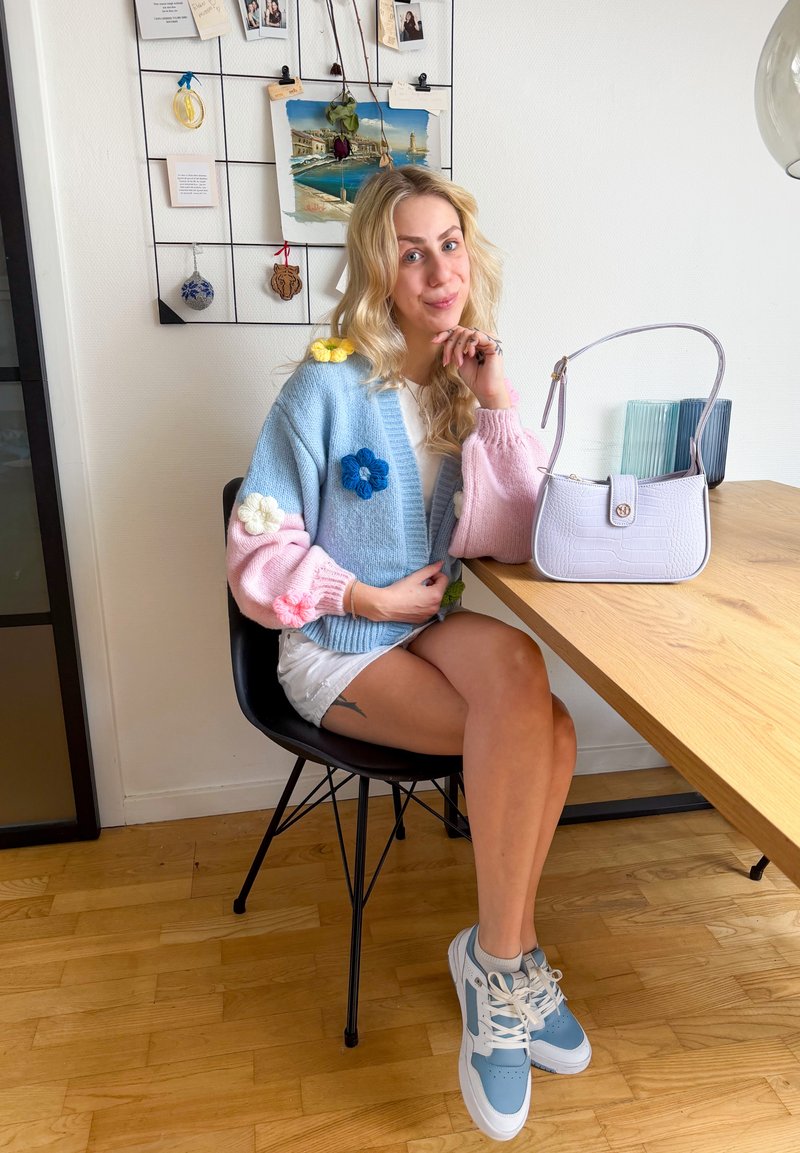 Ung kvinde med langt blondt hår iført en flerfarvet blomstret sweater, hvide shorts og sneakers, sidder ved et træbord med en hvid håndtaske.