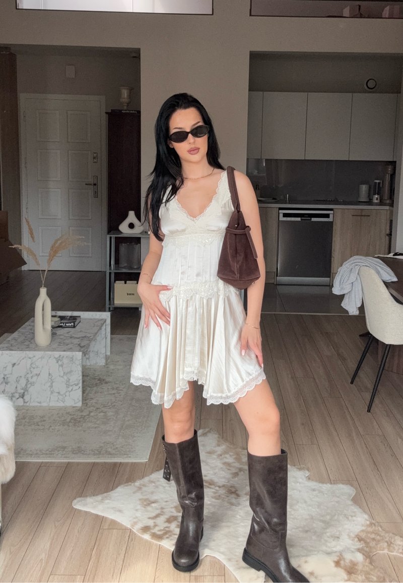 Donna in un vestito bianco con pizzo, stivali neri fino al ginocchio e occhiali da sole, si trova in un soggiorno moderno con pavimento in legno e decorazioni neutre.
