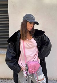 Femme portant une casquette grise, une veste noire oversize, un haut rose, un pantalon gris déchiré et un sac bandoulière rose, debout contre un mur clair.