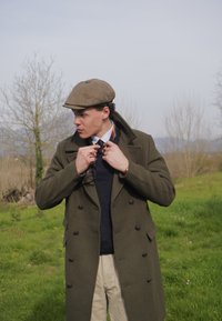 Olivovo zelený dvojradový kabát, námornícky sveter, vzorová kravata, béžové nohavice. Šedý plochý klobúk, drží okuliare, vonku s zeleňou.