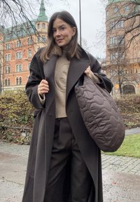 Brązowa pikowana torba z sześciokątnym wzorem, kremowy sweter z tłoczonym motywem, czarny długi płaszcz i ciemne spodnie na zewnątrz.