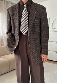 Pruun houndstooth mustriga bleiser suure lõppäisuga ja kahe nupuga, koos ühtlase pruuni särgi ja helesinise ja pruuni triibulise lipsuga.