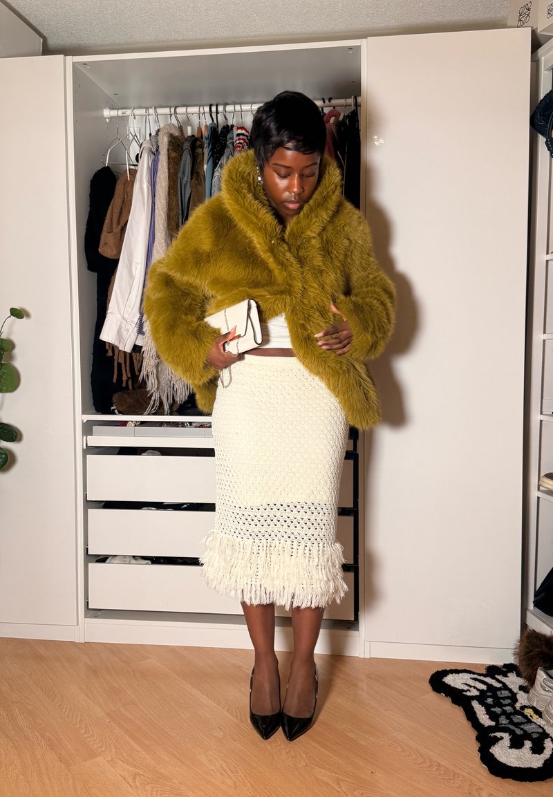Žena v hořčicovém kožichu z umělé kožešiny, bílé pletené sukní s třásněmi, černých podpatcích, držící bílou psaníčko, stojící před otevřenou skříní.