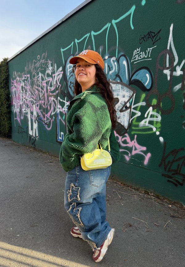 Joven con gorra marrón, chaqueta de forro polar verde, vaqueros con parches y zapatillas, de pie junto a una pared verde cubierta de grafitis, sonriendo a la cámara.
