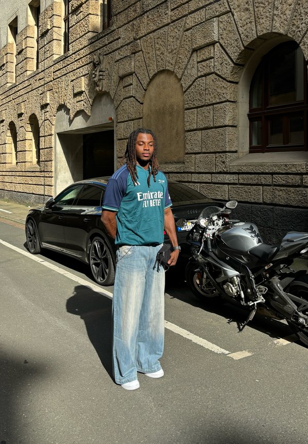 Persona vestita con una maglia sportiva blu-verde con testo bianco, jeans larghi sbiaditi e sneakers bianche, in piedi accanto a un'auto nera e una motocicletta.