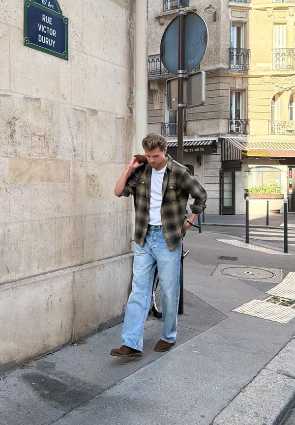 Joven con camisa de cuadros y vaqueros caminando por una calle de París cerca del cartel de Rue Victor Duruy y señales de tráfico.