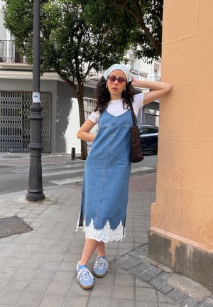 Mujer con gafas de sol, camiseta blanca, vestido vaquero azul con encaje blanco, zapatillas azules y pañuelo blanco en la cabeza, de pie en la acera de la ciudad.