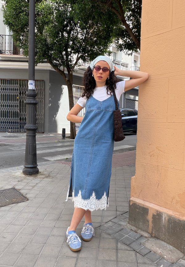 Mujer con gafas de sol, camiseta blanca, vestido vaquero azul con encaje blanco, zapatillas azules y pañuelo blanco en la cabeza, de pie en la acera de la ciudad.