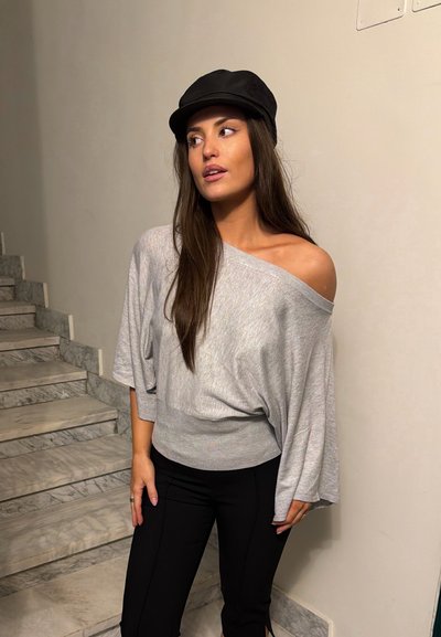 Joven mujer con gorra negra, suéter gris holgado sin hombros y pantalones negros, está de pie cerca de unas escaleras de mármol, mirando hacia su izquierda.