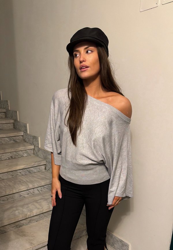 Ung kvinde iført en sort kasket, grå løs sweater med bare skuldre og sorte bukser står nær marmortrapper og ser til venstre.