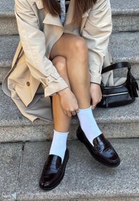 Kvinne i beige trenchcoat som sitter på steintrappen og justerer hvite sokker brukt med mørkebrune loafers, med en svart veske ved siden av seg.