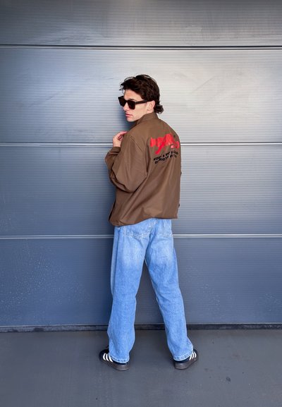 Chaqueta marrón oversized con texto gráfico rojo en la espalda, combinada con jeans baggy de color azul claro y sandalias negras con rayas blancas.