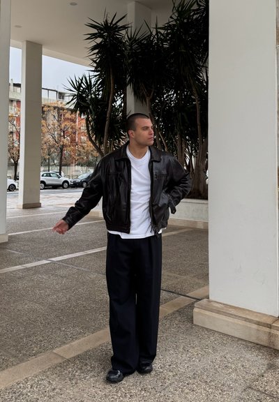 Chaqueta de cuero marrón sobre una camiseta blanca, combinada con pantalones negros holgados y zapatos negros. De pie sobre una superficie exterior texturizada.