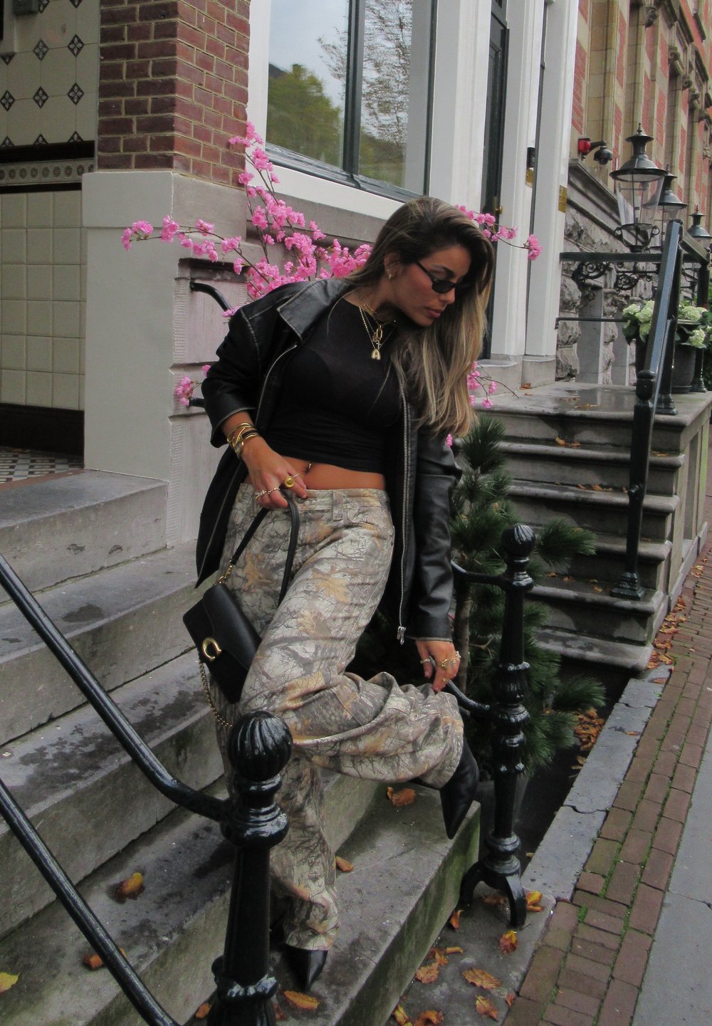 Czarna skórzana kurtka, dopasowany czarny top, spodnie w kamuflażu oraz czarne buty. Stoi na schodach z różowymi kwiatami i ceglaną ścianą w tle.