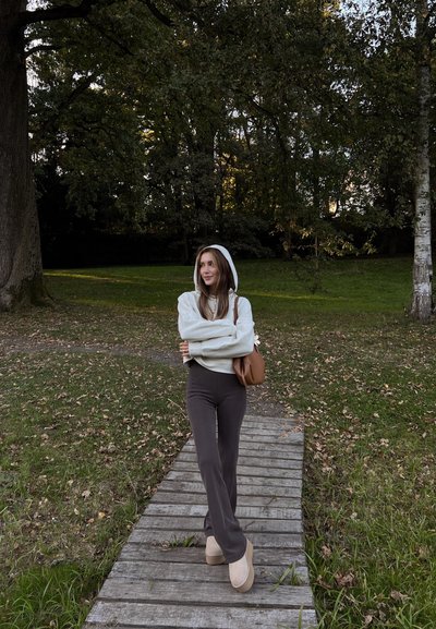Sudadera con capucha gris claro, pantalones de campana gris oscuro, zapatos de plataforma beige, bolso marrón; de pie sobre un camino de madera en un área con hierba.