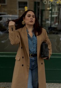 Manteau trench en double-breasted camel porté sur une chemise en denim clair et un jean taille haute. Ceinture noire et pochette. Boucles d'oreilles en or avec des créoles comme détail d'accent.