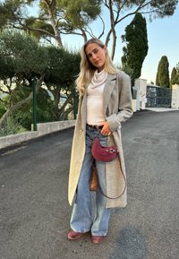Manteau en laine gris clair, pull à col bénitier crème, et jean bleu à jambes larges. Sac à main bordeaux avec ferrures argentées et chaussures assorties. Fond naturel.