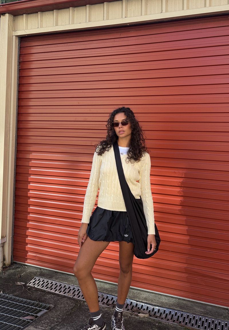 Femme aux cheveux bouclés portant des lunettes de soleil, un pull crème, un short noir et des baskets, debout devant une porte de garage métallique rouge fermée.
