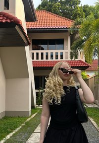 Păr blond, ondulat, cu un top negru fără mâneci și fustă, ținând ochelari de soare. Fundalul prezintă o clădire bej cu acoperișuri de țiglă roșie și vegetație.