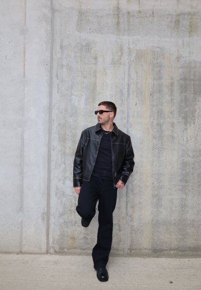 Hombre con chaqueta de cuero negra, pantalones negros y gafas de sol se apoya en una pared de hormigón gris, levantando una pierna y mirando de lado.