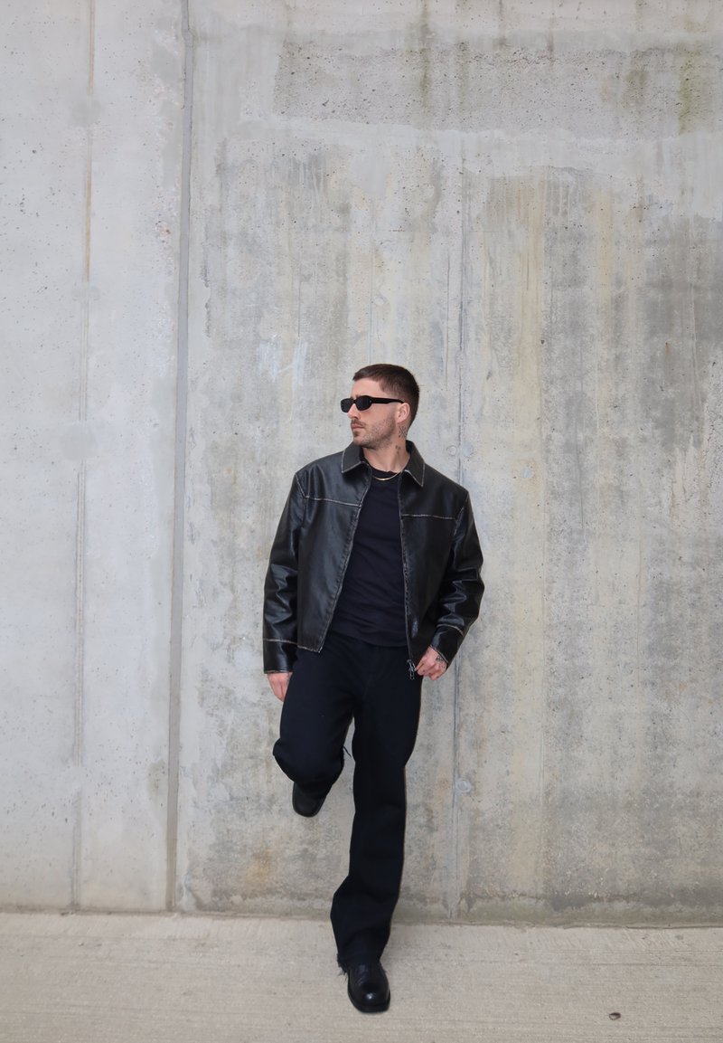 Man draagt een zwarte leren jas, zwarte broek en zonnebril, leunt tegen een grijze betonnen muur, tilt één been op en kijkt opzij.