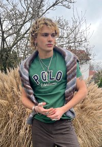 Jeune personne aux cheveux bouclés blonds portant une chemise verte Polo Ralph Lauren et des accessoires, se tenant en plein air près d'herbes sèches et d'arbres dénudés.