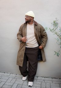 Palton lung maro, pulover bej, pantaloni maro în carouri, teniși albi și căciulă galben deschis; stând lângă un perete simplu cu vegetație.