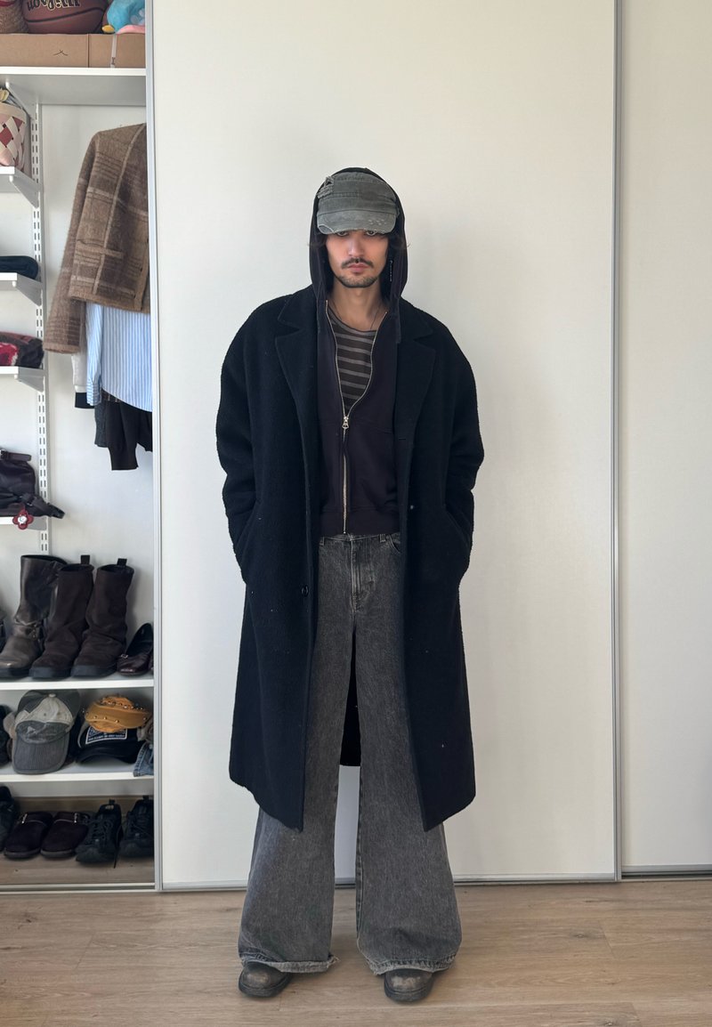 Mees, kes kannab musta pika mantlit, musta kapuutsi, triibulist särki, laiu halli teksapükse ja kulunud halli nokamütsi, seisab käed taskutes toas, kus on riiulid.